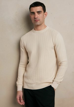 Sweter w kolorze kremowym z dzianiny, z teksturowanym wzorem zygzaków, okrągłym dekoltem, ściągaczami przy mankietach i u dołu, w zestawieniu z ciemnymi spodniami.