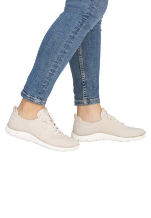 Stopy w beżowych knitowanych tenisówkach z białymi podeszwami i niebieskich obcisłych dżinsach na białym tle