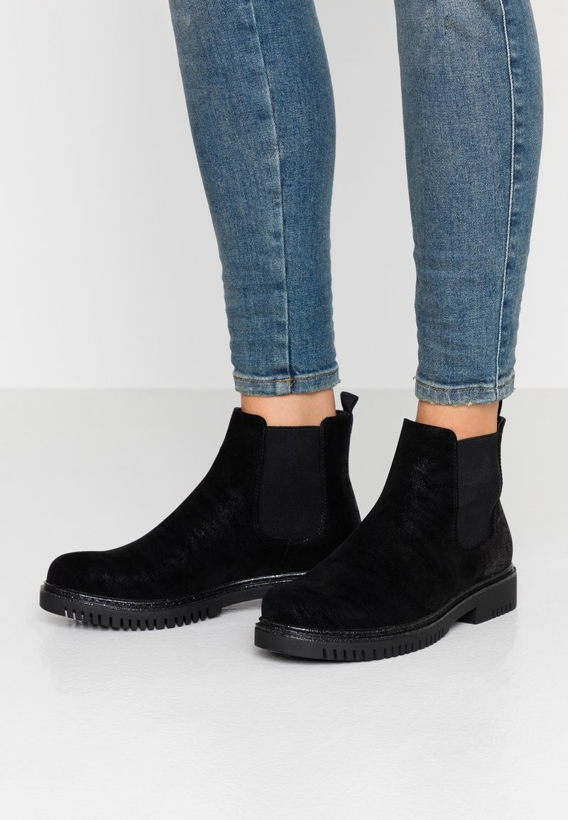 Personne portant un jean slim bleu et des bottines Chelsea en daim noir avec des semelles en caoutchouc rainurées, debout sur une surface blanche lisse.