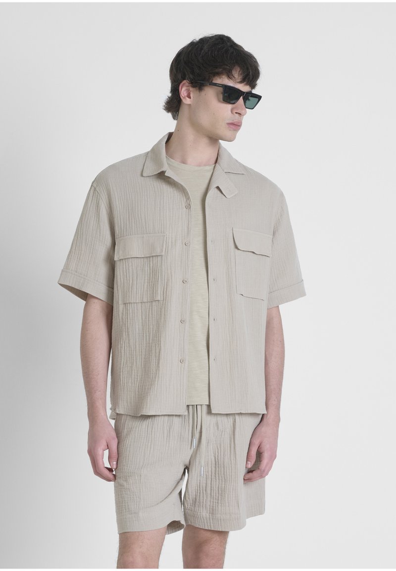 Camicia beige a maniche corte con texture e due tasche frontali, abbinata a pantaloni corti coordinati. Il modello indossa occhiali da sole neri.