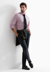 Camisa rosa claro, corbata de cuadros oscuros, suéter verde atado a la cintura, pantalones grises, zapatos negros. Tela suave y corte entallado con tatuajes visibles.