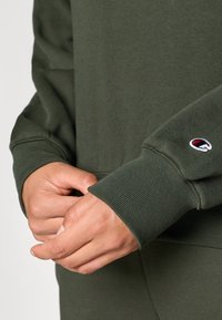 Khaki zöld pulóver bordázott mandzsettákkal és aljjal, bal mandzsettáján kör alakú hímzett logóval. Puha, texturált anyag.