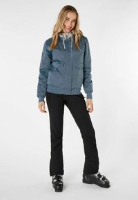 Giacca bomber azzurra con due tasche frontali, polsini elasticizzati e cappuccio regolabile. Indossata con pantaloni da sci neri e scarponi da sci.