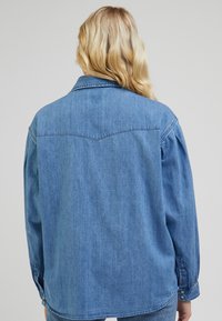 Blaue Jeanshemd mit spitzem Kragen, langen Ärmeln und geknöpften Manschetten. Verfügt über eine Schulternaht und einen geraden Saum.