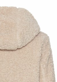 Manteau en beige clair, pelucheux, avec une grande capuche repliée. Présente une texture douce et un design décontracté. Convient pour une tenue relaxante et confortable.