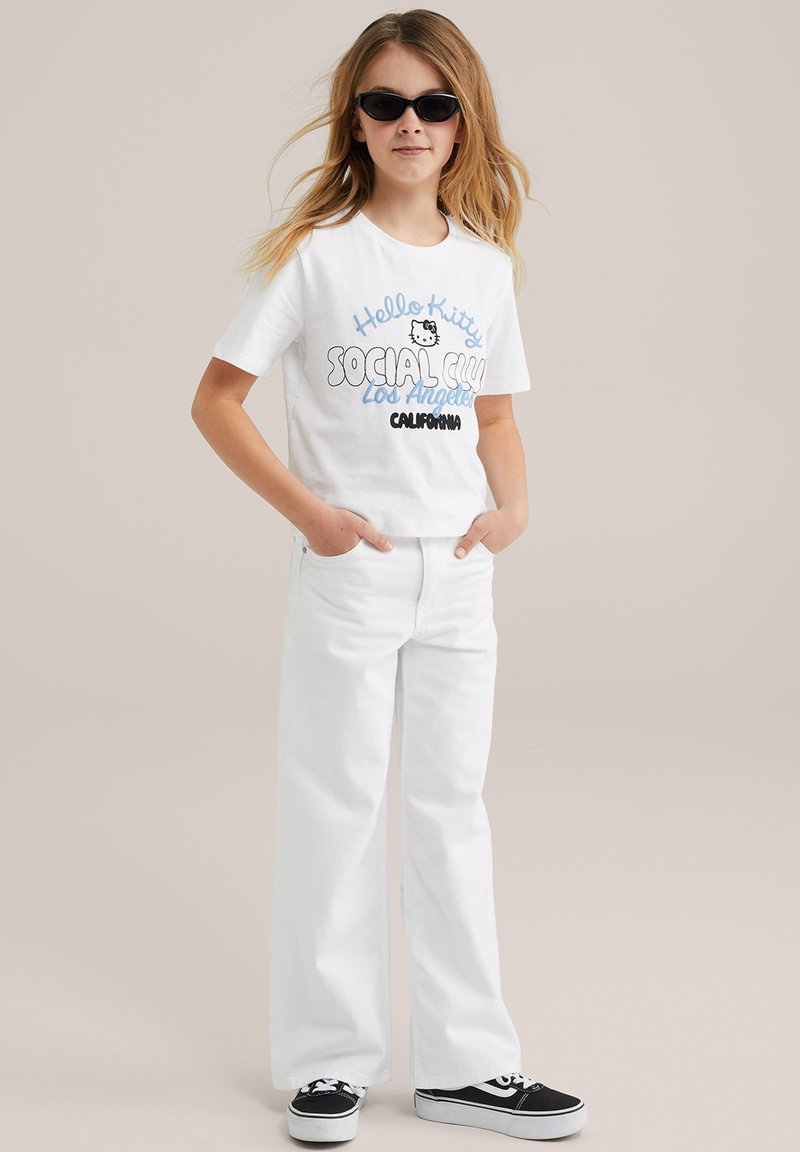 T-shirt blanco corto con un gráfico de "Hello Kitty", combinado con jeans anchos blancos y zapatillas de lona negras. Se llevan gafas de sol negras.