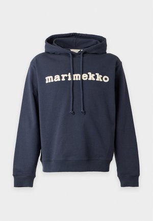 Joven con sudadera azul marino con texto "marimekko", vaqueros azules y zapatillas blancas, de pie frente a un fondo liso y claro.