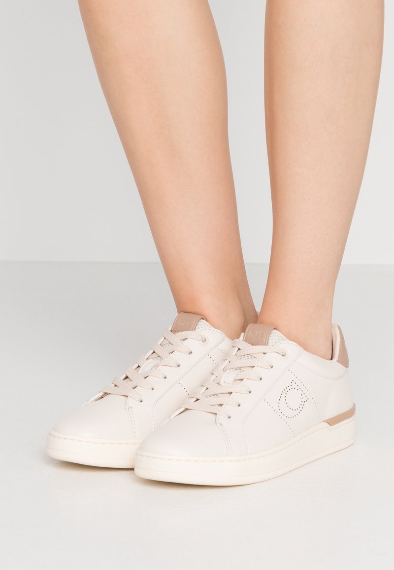 Baskets basses blanches avec des accents beiges et des lacets, portées sur des jambes nues, debout sur un sol gris clair uni.