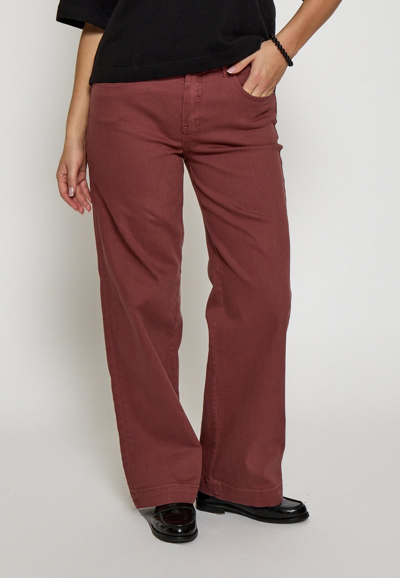 Platās kājas bikses pelēkā vīna krāsā, izgatavotas no izturīga kokvilnas auduma. Rūpīgi izstrādātas ar priekšējām kabatām un brīvu piegriezumu. Melni apavi papildina izskatu.