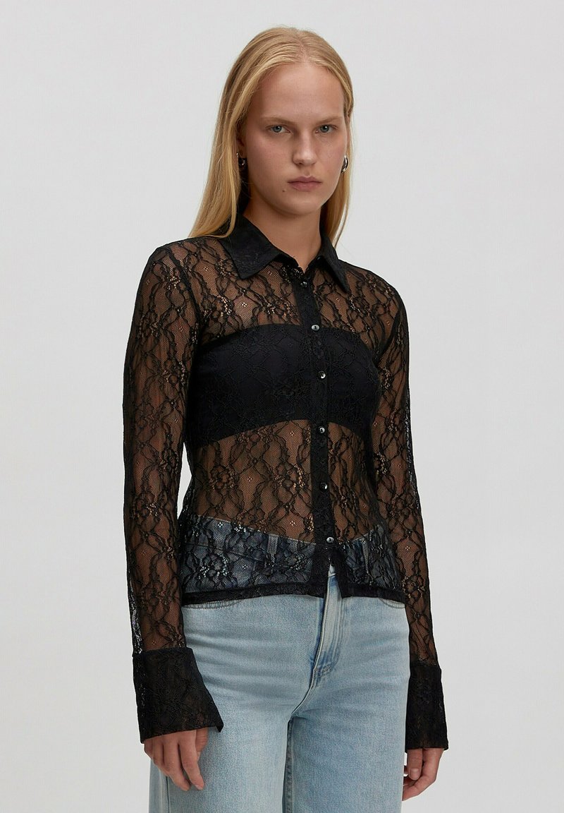 Jonge vrouw die een doorschijnende zwarte kanten knoopbloes over een zwarte bandeau-top draagt, gecombineerd met lichtblauwe high-waisted jeans, staande in een neutrale houding.