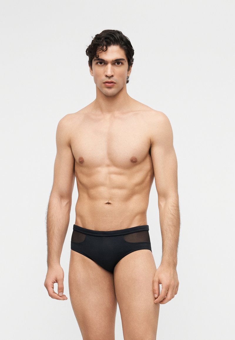 Άνδρας μοντέλο με αδύνατο σώμα και σκούρα μαλλιά, φορώντας μαύρα εσώρουχα με διαφανείς πλέξεις, στέκεται μπροστά σε απλό ανοιχτό γκρι φόντο, κοιτάζοντας προς τα εμπρός.