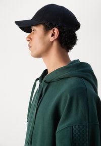 Grønn zip-up hettejakke med en teksturert skulderpanel, kombinert med en svart caps. Hettejakken har snor og mykt stoff.