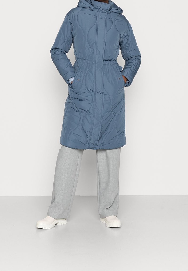 Abrigo acolchado azul claro con capucha desmontable, cintura con cordón, dos bolsillos laterales y textura acolchada, combinado con pantalones gris claro y zapatos blancos.