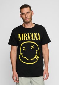 Sort bomulds-T-shirt med stor gul "NIRVANA" tekst og et smilende ansigt med krydsede øjne, præsenteret på en ensartet baggrund.