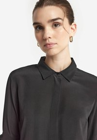 Camicia nera in satin con colletto classico, chiusura a bottoni e un leggero lucido. Tessuto liscio con silhouette diritta e maniche lunghe.