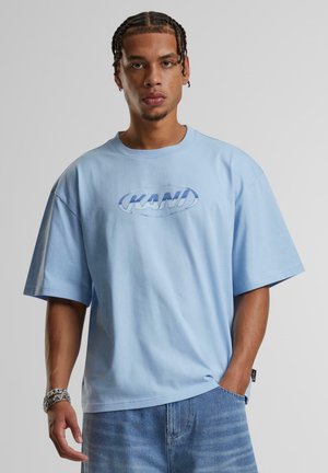Camiseta de algodón oversized de color azul claro con cuello redondo y mangas cortas; presenta una impresión de logo azul en el pecho; combinada con pantalones cortos de mezclilla.