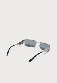 Ochelari de soare metalici rectangulari cu lentile gri închis, cadru argintiu și brațe negre. Prezintă un branding minim pe lateral.