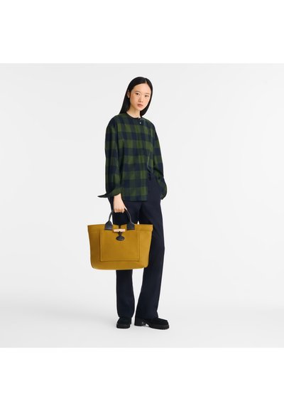 Yellow tote bag with black accents, structured shape, and exterior pocket. Model wears green and navy plaid shirt and navy trousers.