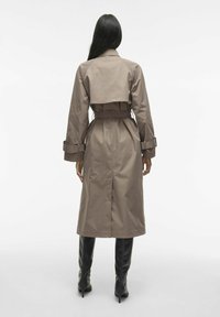 Trench coat beige con vita a cintura, design a manica raglan e dettagli sui polsini. Ha una fessura sul retro per facilitare i movimenti ed è lungo fino al ginocchio.