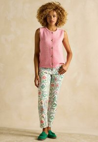 Gilet smanicato rosa in lana con bottoni dorati, abbinato a pantaloni floreali a motivi verdi, bianchi e rosa. Scarpe mocassino in camoscio verde.