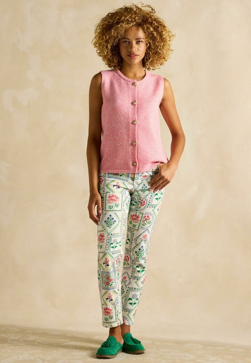 Gilet smanicato rosa in lana con bottoni dorati, abbinato a pantaloni floreali a motivi verdi, bianchi e rosa. Scarpe mocassino in camoscio verde.