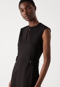 Black dress with a high neckline, cap sleeves, and a keyhole detail. Features side pockets with fabric belts and silver accents. Smooth texture.