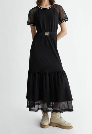 Sort kjole med design af netærmer, flared lagdelt bund og et dekorativt bælte. Sæt sammen med beige snørebodstøvler.