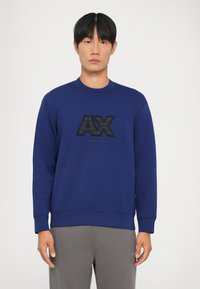 Il pullover blu con collo tondo presenta un prominente logo "AX" nero e del testo sulla parte anteriore. Il materiale sembra morbido con una texture liscia.