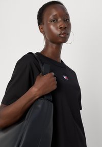 Tricou negru din bumbac cu un mic logo roșu, alb și albastru. Modelul ține o geantă din piele neagră, cu o textură netedă și o formă rotundă.