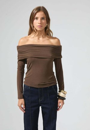 Femme portant un haut brun à épaules nues à manches longues et un jean bleu foncé, accessoirisée avec de grands bracelets au poignet droit.