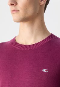 Lilla strikket genser med ribbet rund hals og en liten logo-patch i rødt, hvitt og blått på venstre side av brystet.