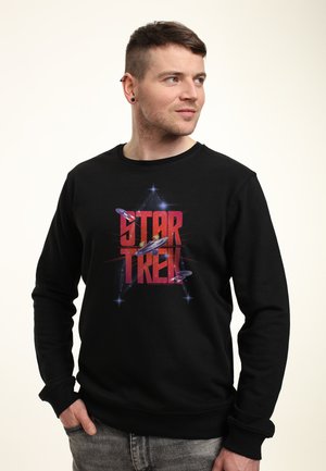 Uomo che indossa una felpa nera di Star Trek con grafiche di astronavi, mani nelle tasche, guarda verso destra su uno sfondo chiaro e semplice.