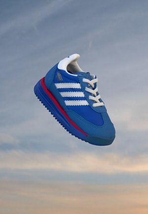 Sininen Adidas-urheilukenkä, jossa valkoiset raidat ja nauhat sekä punainen raita pohjassa, leijailee pilvistä taivasta vasten.