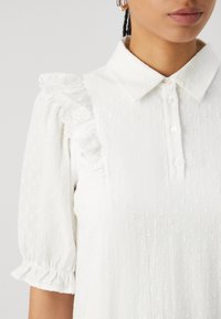 Vit blus med korta puffärmar, spetsdetaljer på axlarna, texturerad fabric och en krage med knappslå.