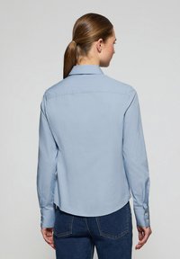Jasnoniebieska koszula z długim rękawem, z wywijanym kołnierzykiem, gładką fakturą i zaokrąglonym dołem. Posiada zapięcie na guziki przy mankietach oraz dopasowany krój.