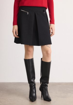 Kobieta nosząca czarne, skórzane kozaki do kolan, czarną plisowaną spódnicę z kieszenią na zamek oraz czerwony, długi rękaw, stojąca na szarej podłodze.