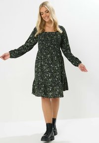 Musta ja vihreä kukallinen mekko, jossa on neliön muotoinen pääntie, pitkät puff-hihat, rypytetty yläosa ja kerroksellinen hame, yhdistettynä mustiin nauhakengiin.