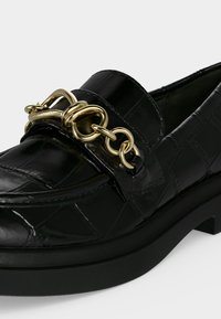 Mocassin en cuir noir avec motif crocodile et chaîne dorée décorative sur le dessus.