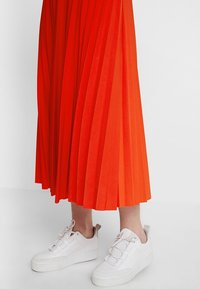 Falda plisada hasta el tobillo en un vibrante color naranja, combinada con zapatillas blancas que presentan una suela texturizada y múltiples ojales para los cordones.