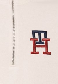 Polo s půlzipem v krémové barvě, který má na hrudi vyšitý červený a námořnický logo "TH" umístěné v centru, s hladkou texturou.