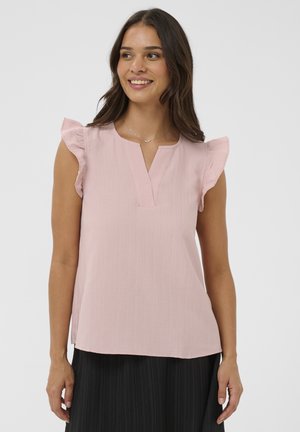 Nő világos rózsaszín ujjatlan blúzzal, fodros vállal és fekete pliszírozott szoknyával, mosolyog és oldalra néz.