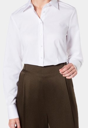 Persona che indossa una camicia bianca abbottonata e pantaloni marroni a vita alta, mostrata dalle spalle alle cosce su uno sfondo neutro.