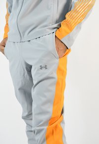 Chándal atlético gris con rayas laterales naranjas, hecho de un tejido suave y ligero. Logo notable en la pierna y diseño de chaqueta con cremallera.