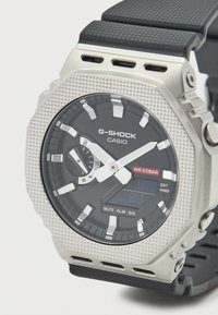 Ρολόι G-SHOCK με θήκη από ανοξείδωτο ατσάλι, μαύρο λουράκι από καουτσούκ, μαύρο καντράν με λευκές και κόκκινες λεπτομέρειες, και υφή στη θήκη. Αδιάβροχο έως 20 bar.