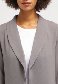Šedý, lehký blazer s šálovým límcem, texturovanou tkaninou a otevřeným předním designem. Nositelný přes bílou košili.