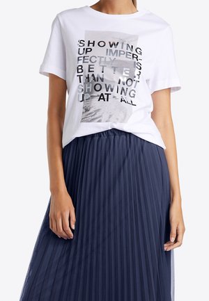 Kobieta w białej koszulce z czarnym i szarym tekstem oraz granatowej plisowanej spódnicy, pokazana od ramion do połowy uda.