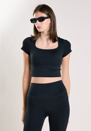 Donna che indossa un crop top nero aderente, leggings a vita alta e occhiali da sole neri rettangolari su uno sfondo chiaro e uniforme.