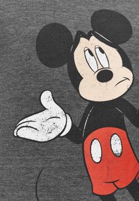 Personnage de dessin animé classique avec de grandes oreilles rondes, main gantée blanche tendue, short rouge avec boutons blancs, sur fond de tissu gris.