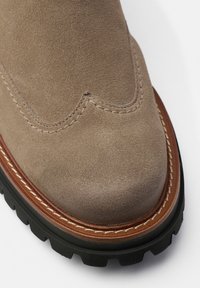 Botte avec une tige en daim taupe, présentant un design festonné, des coutures contrastées et une semelle en caoutchouc noire avec une adhérence texturée.