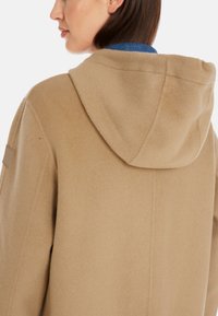 Warmer beige Kapuzenmantel aus weichem Stoff, mit glatter Textur und einem kleinen Logopatch an der Schulter, sichtbare Nahtdetails.